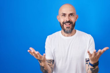 Mavi arka planda dikilen İspanyol dövmeli adam güler yüzlü, dostça kollar açık, olumlu ve kendinden emin selamlar. 