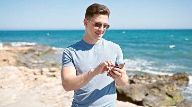 Genç İspanyol turist deniz kenarında akıllı telefon kullanarak gülümsüyor.