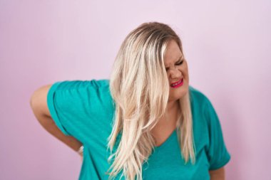 Beyaz tenli, uzun boylu kadın pembe arka planda duruyor sırt ağrısı çekiyor, elleriyle dokunuyor, kas ağrısı çekiyor. 