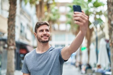 Kafkasyalı genç adam gülümsüyor. Sokakta akıllı telefonun yanında özçekim yapıyor.