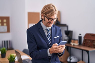 Genç sarışın iş adamı ofisteki akıllı telefonu kullanarak gülümsüyor.