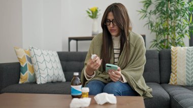 Genç, güzel, İspanyol bir kadın elinde ilaç şişesiyle evde akıllı telefon kullanıyor.