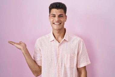 Young hispanic man standing over pink background smiling cheerful presenting and pointing with palm of hand looking at the camera. 