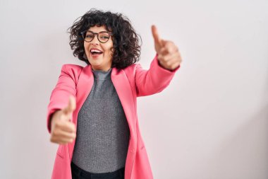 Kıvırcık saçlı İspanyol kadın izole edilmiş arka planda duruyor. Elleriyle olumlu jest yapıyor, gülümsüyor ve başarı için mutlu. kazanan jest. 