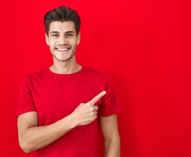 Genç İspanyol bir adam kırmızı arka planda neşeli bir şekilde ayakta duruyor. Yüzünde mutlu ve doğal bir ifadeyle elleri ve parmaklarıyla işaret eden bir gülümseme var. 
