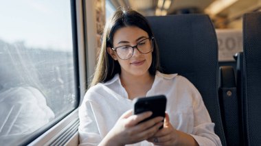 Genç, güzel, İspanyol bir kadın gülümsüyor. Akıllı telefon kullanıyor. Trende oturuyor.