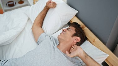 Young hispanic man waking up stretching arms at bedroom