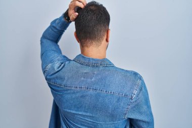 Genç İspanyol adam mavi arka planda dikiliyor. Elleri kafasında şüpheyi düşünüyor. 