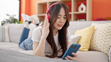 Genç Çinli kadın akıllı telefon kullanıyor kulaklık takıp evdeki kanepede yatıyor.