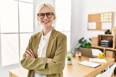 Genç, sarışın, iş kadını, gülümseyen, kendine güvenen, kollarını kavuşturup ofiste el kol kola duran.