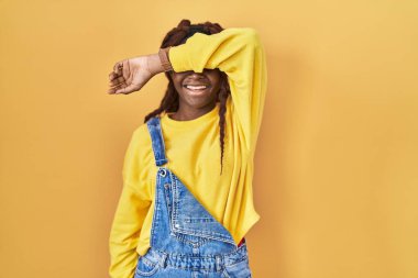 Sarı arka planda dikilen Afrikalı kadın kollarıyla gözlerini kapatıyor neşeli ve komik gülümsüyor. kör kavram. 
