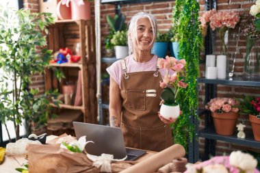 Middle age grey-haired woman florist using laptop holding plant at florist