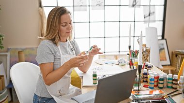Güzel sarışın sanatçı, tutkuyla sanat yaratıyor, elinde renkli bir tüp tutuyor, canlı stüdyosunda dizüstü bilgisayarına takılı..
