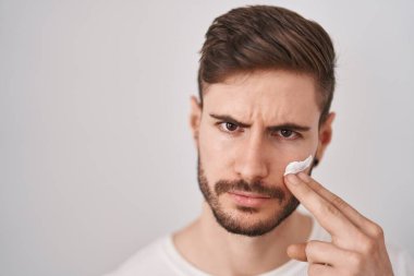 Sakallı İspanyol bir adam nemlendirici yüz kremi kullanıyor şüpheci ve gergin, sorun yüzünden kaşlarını çatıyor. negatif kişi. 