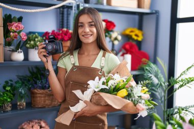Çiçekçide çalışan esmer bir kadın elinde kamerayla mutlu ve havalı bir gülümsemeyle gülümsüyor. diş gösterme. 
