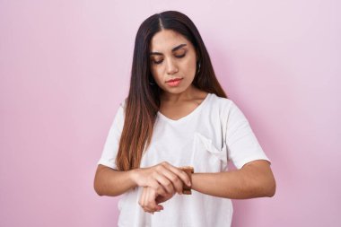 Pembe arka planda duran genç Arap kadın kol saatini kontrol ediyor, rahat ve kendinden emin. 