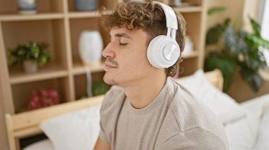 Genç, yakışıklı İspanyol adam rahat bir şekilde rahatlıyor, neşe içinde eğlenceli müzikler dinliyor, sade yatak odasında rahat bir yatakta oturuyor, mutluluğu ve güveni somutlaştırıyor ve sabahın tadını çıkarıyor.