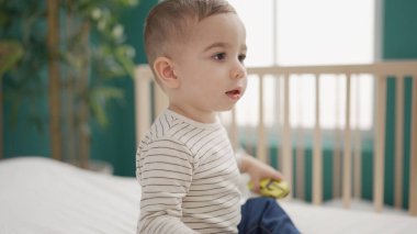 Sevimli çocuk yatakta oturmuş, yüzünde ciddi bir ifadeyle yatak odasında araba oyuncağıyla oynuyor.