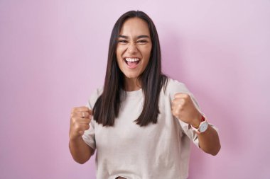 Pembe arka planda duran genç İspanyol kadın mutlu ve heyecanlı bir şekilde kollarını kaldırıp, gülümseyip, başarı için çığlık atıyor. kutlama konsepti. 