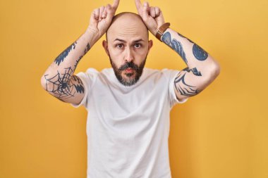 Sarı arka planda dikilen İspanyol dövmeli genç adam parmaklarını boynuzlarının üzerinde gezdirirken komik hareketler yapıyor. 