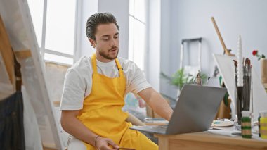 Genç İspanyol ressam, sanat stüdyosunda ciddi yüzlü dizüstü bilgisayar kullanıyor.