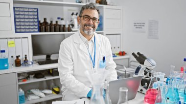 Çekici, gri saçlı, İspanyol asıllı genç bir adam, kendinden emin bir bilim adamı, dolup taşan bir laboratuvarda dizüstü bilgisayarını kullanarak biyoloji analizi yaparken gülümsüyor..