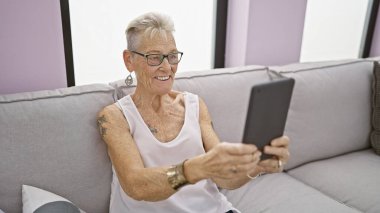 Kendine güveni tam, kısa gri saçlı, rahat koltukta dokunmatik ped kullanan olgun bir kadın. Orta yaşlı bir kadın oturma odasında teknolojinin tadını çıkarıyor..