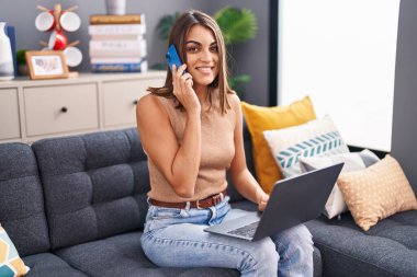 Genç, güzel, İspanyol bir kadın akıllı telefonuyla evde dizüstü bilgisayar kullanıyor.