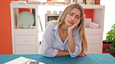 Genç sarışın kadın baş ağrısı çekiyor. Yemek masasında oturuyor.