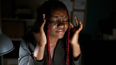 Yorgun afro-amerikan kadın işçi, iş kıyafetleri içinde, örgüler sıkı çekti, bilgisayarla aydınlatılmış çalışma odasında internet stresinde geziniyor, gece boyunca korkunç bir baş ağrısıyla mücadele ediyor.