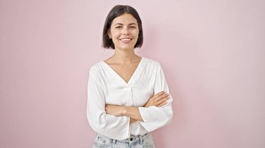 Young beautiful hispanic woman smiling confident standing with arms crossed gesture over isolated pink background