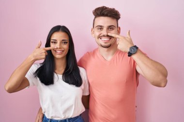 Young hispanic couple standing over pink background pointing with hand finger to face and nose, smiling cheerful. beauty concept 