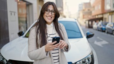 Genç, güzel, İspanyol bir kadın akıllı telefon kullanıyor. Arabasının yanında duruyor.