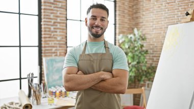 Kollarını kavuşturup sanat stüdyosuna gülümseyen genç Arap sanatçı.