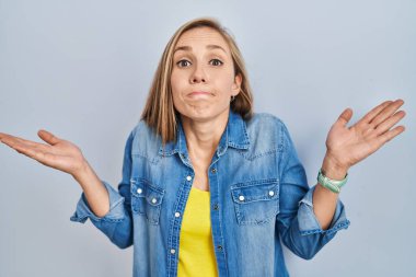 Mavi arka planda duran genç sarışın kadın, elleri ve kolları havada, kafası karışmış bir ifade takınıyor. şüphe kavramı. 