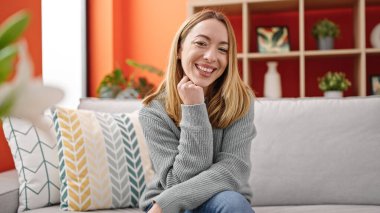 Sarışın genç bir kadın evdeki koltukta kendinden emin bir şekilde oturarak gülümsüyor.