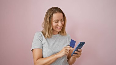 Çekici, kendine güveni tam, pembe arka planda gülümseyen genç bir kadın. Elinde kredi kartı tutarken akıllı telefon kullanıyor. Mutluluk ve finansal bağımsızlık yayıyor.