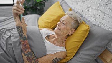 Mutlu orta yaşlı bir kadın, özgüvenle gülümsüyor, rahat bir yatakta dinleniyor, internette mesajlaşıyor. Beyaz saçlı kadın, pijamalı, rahat bir yatak odasında telefonunu kullanıyor..