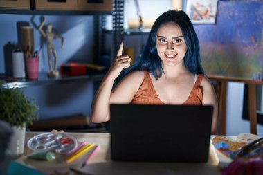 Mavi saçlı genç modern kız gece vakti dizüstü bilgisayarla sanat stüdyosunda oturuyor ve başarılı bir fikir sunuyor. Heyecanlı ve mutlu. Bir numara.. 