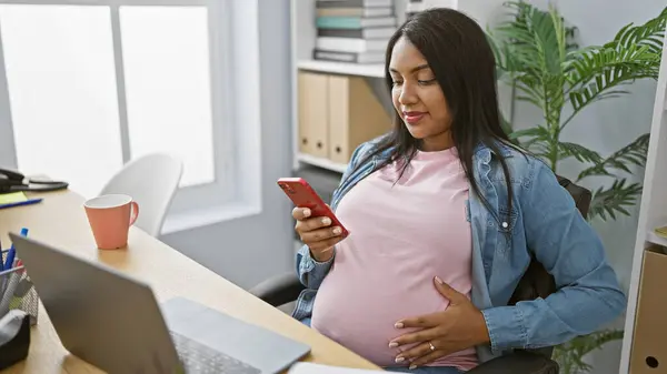 有自信的年轻孕妇 一边在办公室里用智能手机工作一边高兴地摸着肚子 体现了成功的母性 — 图库照片