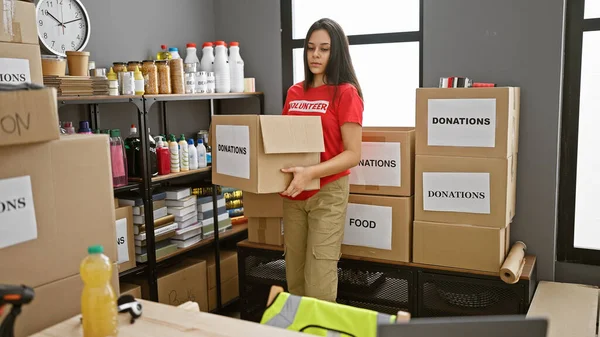 Beautiful young hispanic woman focused on altruism, volunteering at charity center, holding donations package, standing strong in unity, working hard in workplace interior