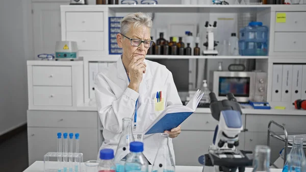 Deep in thought, a grey-haired senior woman scientist studying and reading an intriguing medical book at her laboratory.