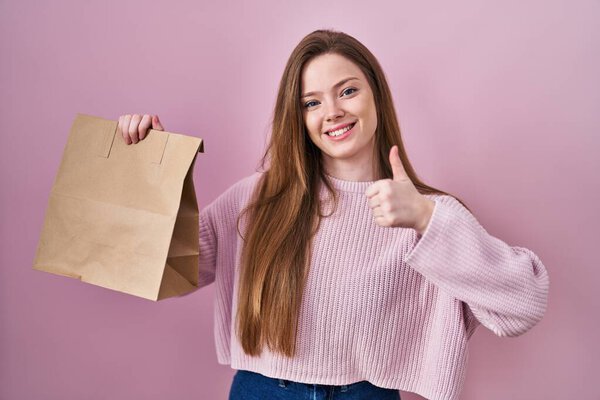 Young caucasian woman holding shopping bag and credit card smiling happy and positive, thumb up doing excellent and approval sign 