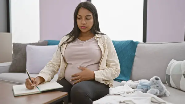 安详的年轻惊慌失措的孕妇爱检查婴儿衣服 边做笔记边舒服地在家里休息 — 图库照片