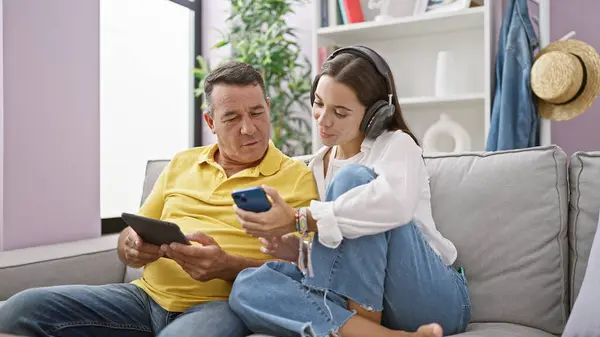 Hispanic father smilingly using smartphone, daughter comfortable on sofa with touchpad, enjoying family tech-time indoors