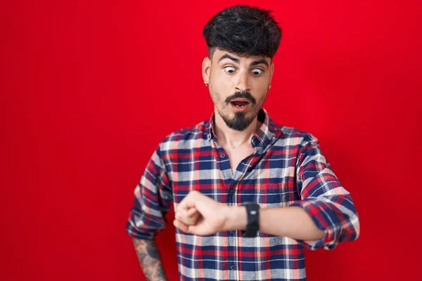 Jovem Hispânico Com Barba Sobre Fundo Vermelho Olhando Para Relógio — Fotografia de Stock