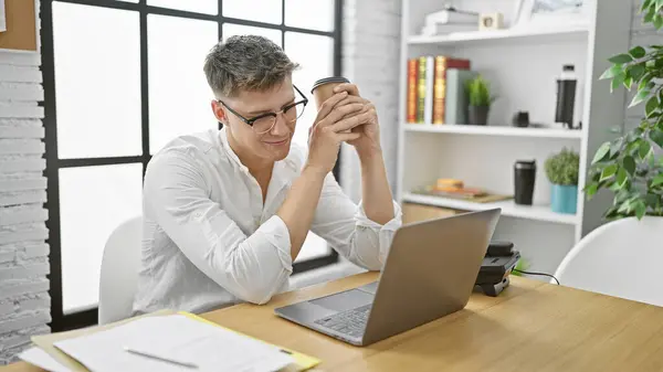 英俊的年轻白种人 自信满满的员工在办公室的办公桌前微笑着 一边享受着早晨的浓缩咖啡 一边与笔记本电脑一起上网 — 图库照片