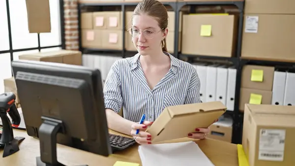 Young blonde woman ecommerce business worker using computer writing on package at office