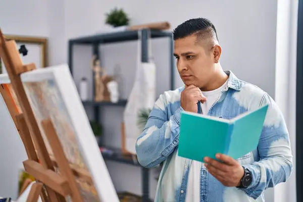 年轻的拉丁艺术家在艺术工作室里带着怀疑的表情看书 — 图库照片