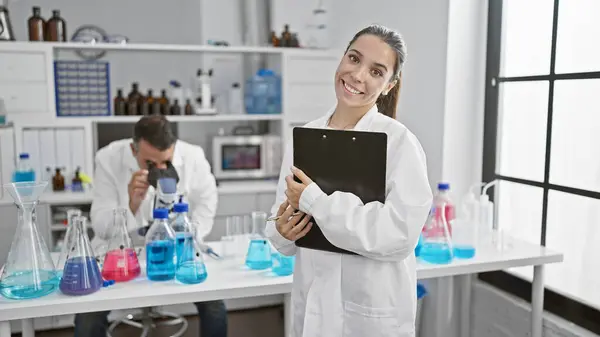 Confident hispanic man and woman scientists holding clipboard, smiling as they discover new medicinal techniques using a microscope in lab